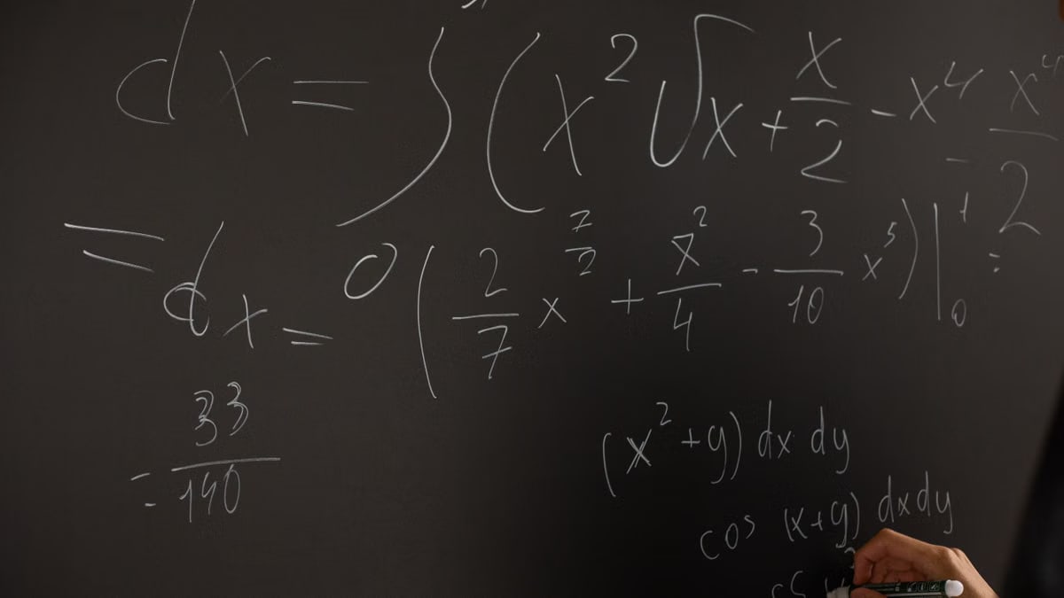 Close-up of hand writing complex math equations on a chalkboard in a classroom setting.
